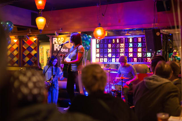 Live music performance at Love Bomb 2017 event, featuring musicians on stage with guitar and drums, colorful lighting, and audience engagement in a vibrant setting.